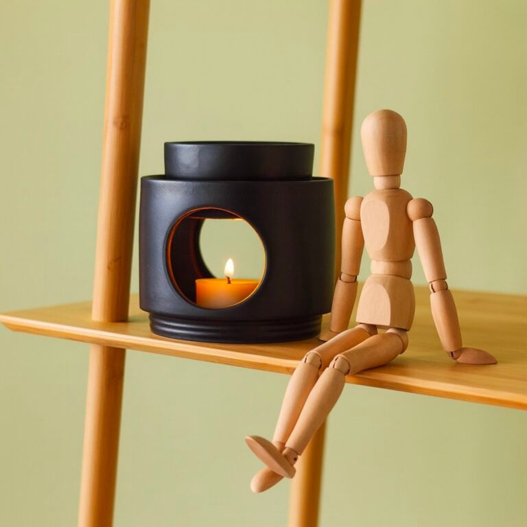 Quemador Noir de cerámica negra SYS Aromas con vela encendida junto a figura articulada de madera